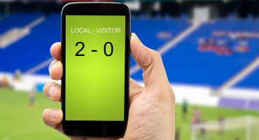 Hand holding a mobile phone displaying a football score, with a stadium in the background