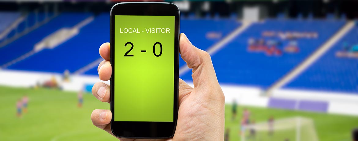 Hand holding a mobile phone displaying a football score, with a stadium in the background